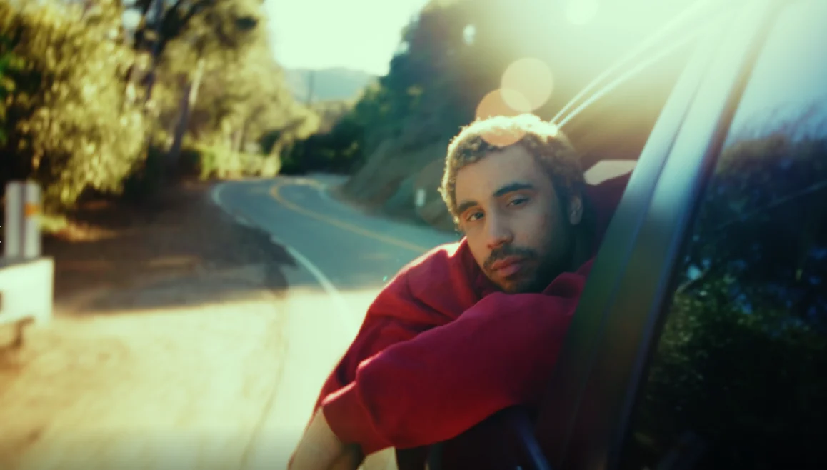A man in a red hoodie leans out of a car window on a winding canyon road in golden hour light