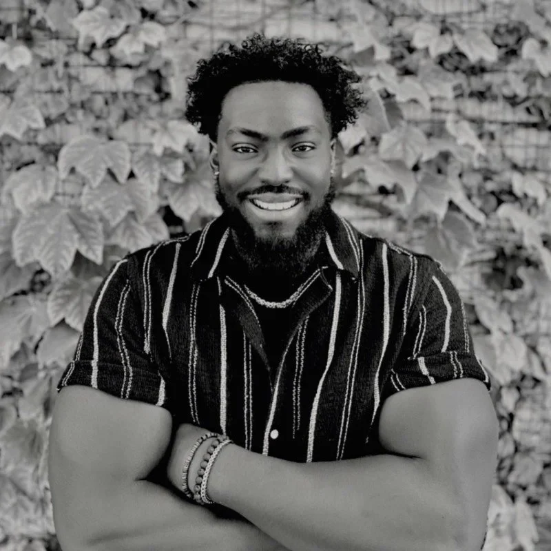 Kameron Gray smiling with arms crossed, in a black and white portrait against an ivy covered wall